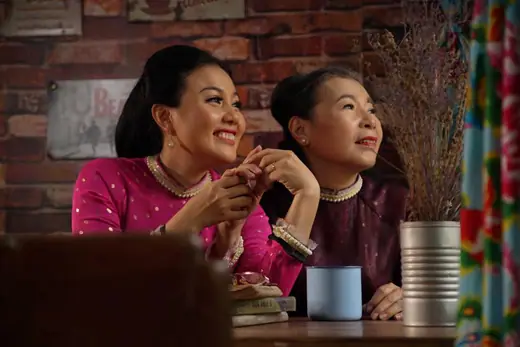 Hai phụ nữ mặc áo dài hồng và tím mận ngồi bên bàn, phía sau là tường gạch.