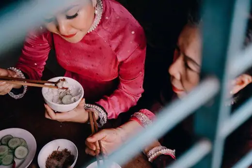 Hai phụ nữ mặc áo dài hồng và tím mận ngồi ăn, chụp từ trên cao qua khung cửa.