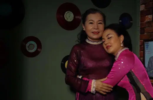 Hai phụ nữ mặc áo dài tím mận và hồng cánh sen ôm nhau trước bức tường treo đĩa nhạc.