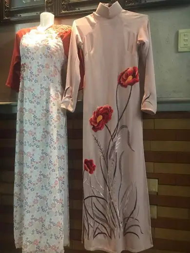 Hai chiếc áo dài trên ma-nơ-canh: một trắng hoa xanh nhạt, một màu be thêu hoa đỏ.