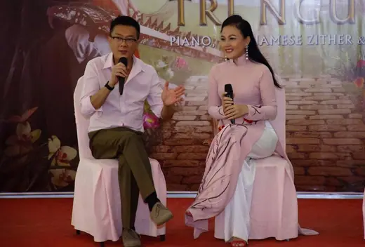Một người đàn ông và một phụ nữ mặc áo dài hồng nhạt ngồi trò chuyện trên sân khấu.