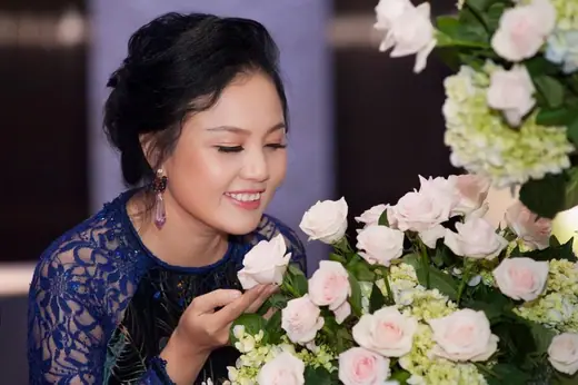 Một phụ nữ mặc áo dài ren xanh tím than cúi ngửi hoa hồng trắng.