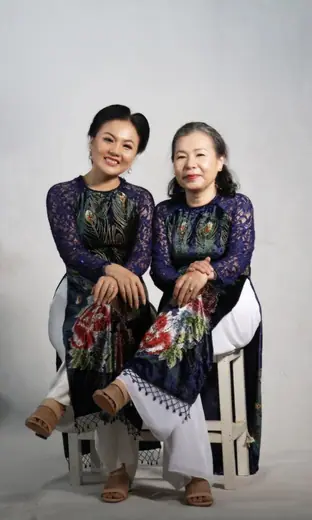 Hai phụ nữ mặc áo dài đôi xanh tím than in chim công ngồi trong phòng chụp.