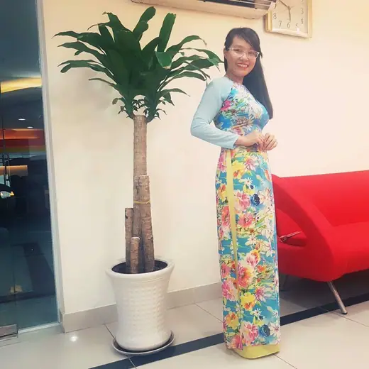 Một phụ nữ mặc áo dài xanh in hoa đứng cạnh chậu cây trong sảnh văn phòng.