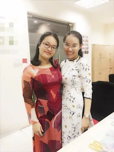 Hai phụ nữ mặc áo dài, một đỏ hoạ tiết và một trắng hoa nhí, trong văn phòng.