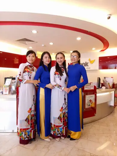 Bốn nữ nhân viên mặc áo dài đồng phục trắng hoạ tiết và xanh dương trong sảnh ngân hàng.