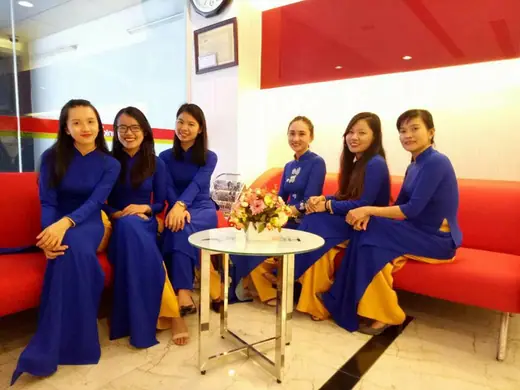 Sáu nữ nhân viên mặc áo dài đồng phục xanh dương ngồi trên sofa đỏ.