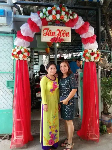 Hai phụ nữ đứng trước cổng hoa đám cưới, một người mặc áo dài vàng in hoa.