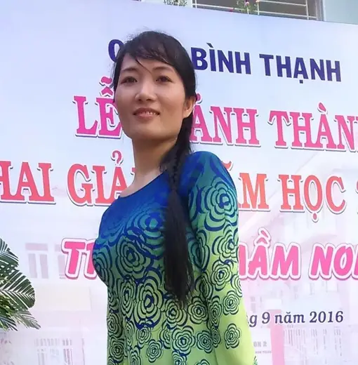 Một phụ nữ mặc áo dài xanh lá phối xanh dương hoạ tiết xoáy đứng trước phông lễ khai giảng.