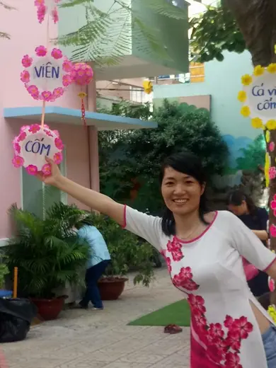 Một phụ nữ mặc áo dài trắng in hoa hồng đứng trong sân trường mầm non.