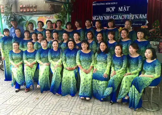 Tập thể cô giáo mặc áo dài xanh lá phối xanh dương chụp trước phông họp mặt ngày Nhà giáo Việt Nam.