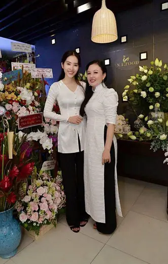 Hai phụ nữ mặc đồ trắng đứng trước cửa tiệm có nhiều lẵng hoa chúc mừng.