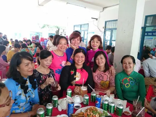 Nhóm phụ nữ mặc áo dài nhiều màu ngồi quanh bàn tiệc trong sân trường.