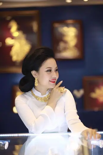 Một phụ nữ mặc áo dài trắng, đeo trang sức vàng, đứng bên quầy kính.