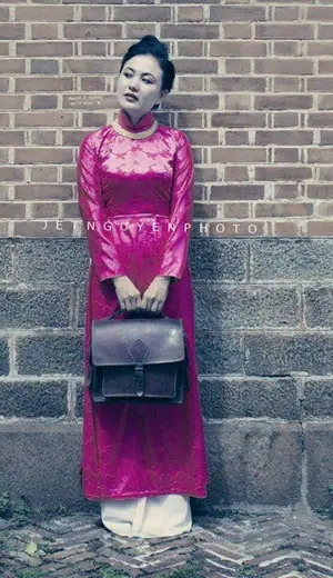 Một phụ nữ mặc áo dài hồng sen kiểu xưa, xách cặp da, đứng trước tường gạch.