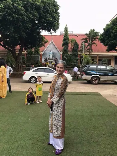 Một cụ bà mặc áo dài gấm đứng trên bãi cỏ trước nhà thờ.