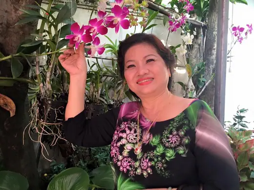 Một phụ nữ mặc áo dài đen thêu hoa đứng dưới giàn lan tím.
