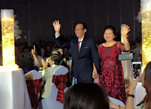 Ông bà sui vẫy tay chào khách khi tiến vào sảnh tiệc cưới, bà mặc áo dài đỏ sẫm.