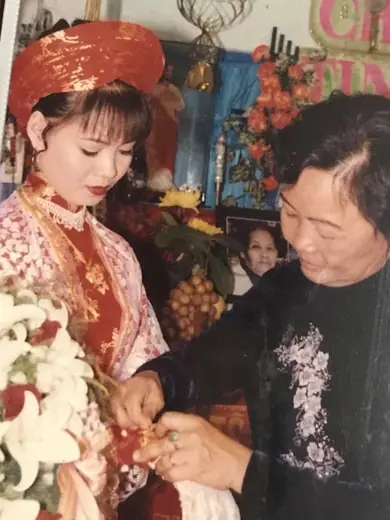 Ảnh cũ: cô dâu mặc áo dài ren đỏ, một phụ nữ mặc áo dài nhung đen thêu hoa đang đeo vòng cho cô dâu.