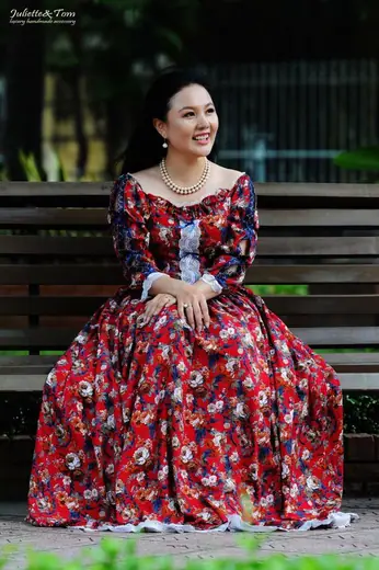 Một phụ nữ mặc váy xoè đỏ in hoa ngồi trên ghế băng trong công viên.