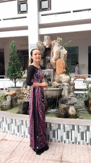 Một cô giáo mặc áo dài lụa tối màu hoạ tiết đứng bên hòn non bộ trong sân trường.