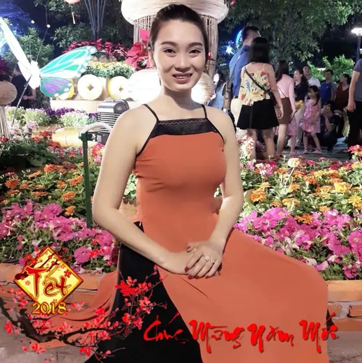 Một phụ nữ mặc áo yếm cam và váy đen ở đường hoa Tết, ảnh có khung chúc mừng năm mới.