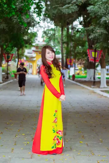 Một cô gái mặc áo dài vàng phối đỏ vẽ hoa đứng trên lối đi rợp bóng cây.