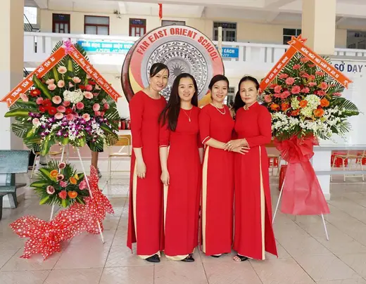 Bốn phụ nữ mặc áo dài đỏ đồng phục đứng giữa các lẵng hoa chúc mừng.