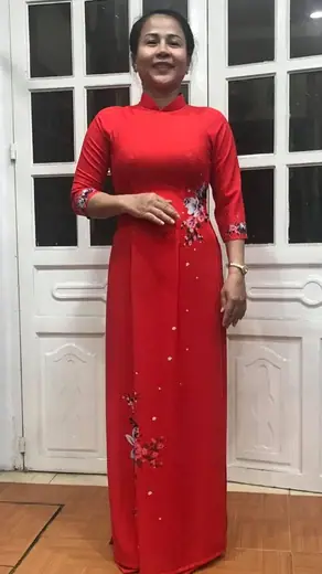 Một phụ nữ mặc áo dài đỏ kết hoa cườm đứng trước cửa.
