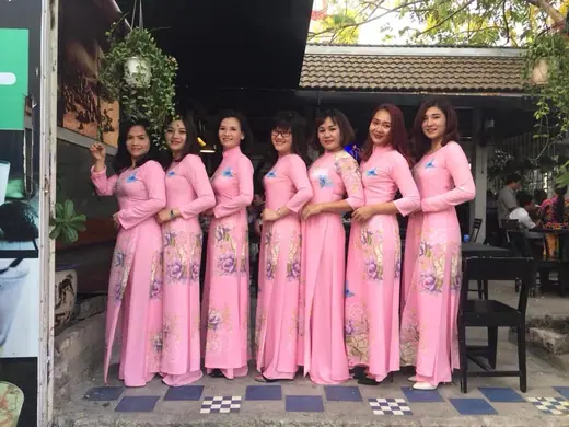 Bảy phụ nữ mặc áo dài đồng phục hồng in hoa đứng thành hàng ở quán cà phê sân vườn.