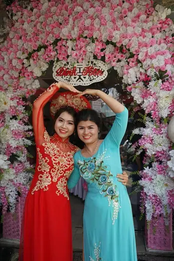 Hai phụ nữ mặc áo dài đỏ thêu vàng và xanh ngọc kết cườm tạo dáng trước cổng hoa.