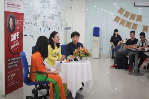 Hai phụ nữ mặc áo dài cam và vàng ngồi trò chuyện trong một phòng họp.