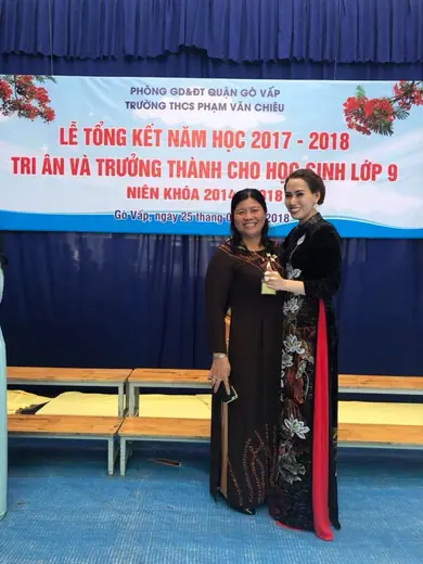 Hai cô giáo mặc áo dài tối màu đứng trước phông lễ tổng kết năm học.