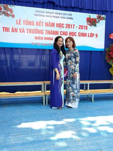 Hai cô giáo mặc áo dài xanh dương và áo dài in hoa trước phông lễ tổng kết năm học.