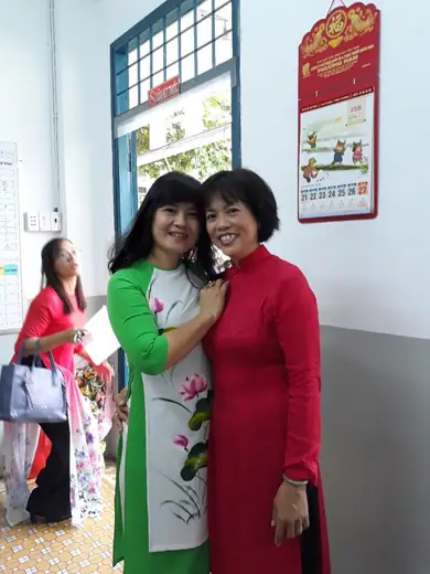 Hai cô giáo, một mặc áo dài trắng vẽ hoa sen phối xanh lá, một mặc áo dài đỏ, đứng ở hành lang trường.