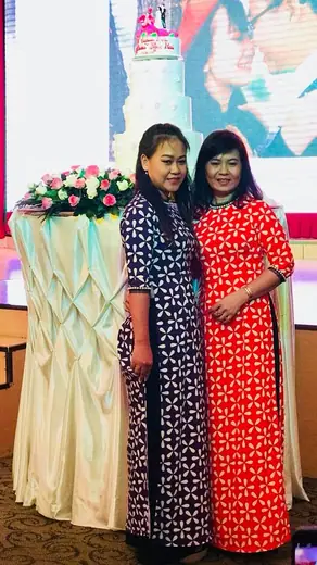 Hai phụ nữ mặc áo dài in hoạ tiết xanh tím than và đỏ đứng cạnh bàn trang trí hoa.
