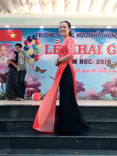 Một cô giáo mặc áo dài đỏ phối đen bước trên sân khấu lễ khai giảng.