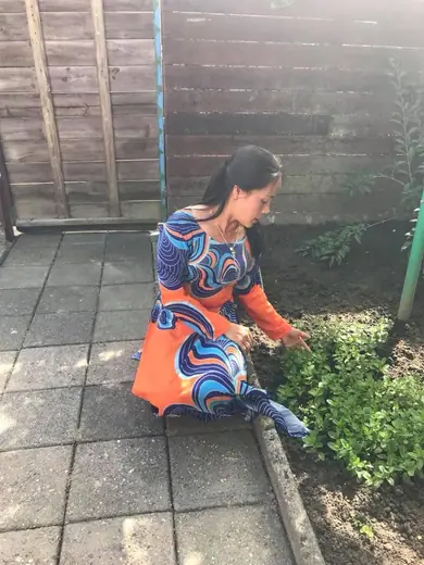 Một phụ nữ mặc áo dài in hoạ tiết xoáy xanh cam ngồi bên luống rau.