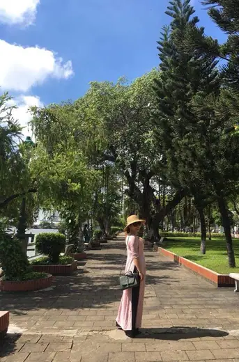 Một cô giáo mặc áo dài hồng nhạt, đội nón, đứng trên lối đi trong công viên.