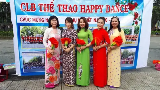 Năm phụ nữ mặc áo dài cầm hoa đứng trước băng rôn của một câu lạc bộ khiêu vũ thể thao.