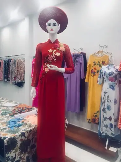 Ma-nơ-canh mặc áo dài đỏ thêu hoa, đội nón, trong tiệm với nhiều áo dài phía sau.