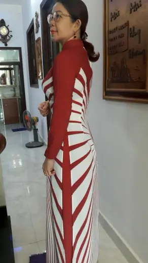 Một phụ nữ mặc áo dài sọc đỏ trắng đứng nghiêng trong nhà.