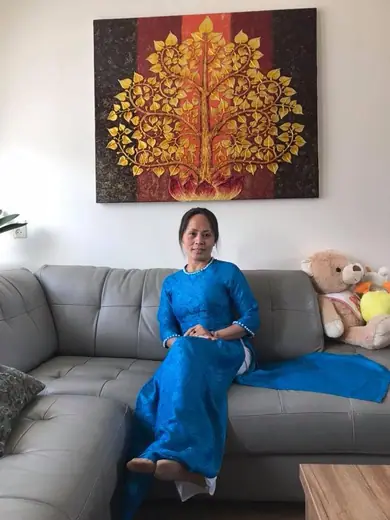Một phụ nữ mặc áo dài gấm xanh ngồi trên sofa xám dưới bức tranh cây vàng.