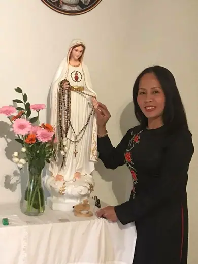 Một phụ nữ mặc áo dài đen thêu hoa hồng đỏ đứng bên tượng Đức Mẹ.