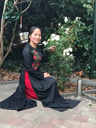 Một phụ nữ mặc áo dài đen thêu hoa hồng đỏ quỳ bên khóm hoa hồng trắng.