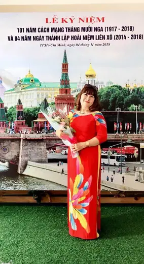 Một phụ nữ mặc áo dài đỏ vẽ mảng màu ôm bó hoa trước phông lễ kỷ niệm.