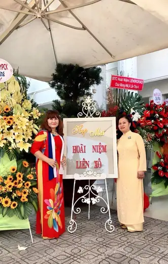 Hai phụ nữ mặc áo dài đứng bên bảng hiệu sự kiện giữa các lẵng hoa.