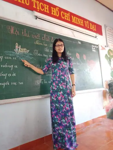 Một cô giáo mặc áo dài lụa in hoa đứng giảng bài bên bảng lớp học.