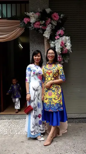 Hai phụ nữ mặc áo dài cách tân, một trắng in hoa và một hoạ tiết cung đình vàng xanh, trước cổng hoa đám cưới.