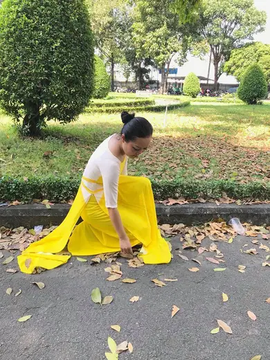 Một cô giáo mặc áo dài vàng phối trắng ngồi trên bãi cỏ đầy lá rụng.